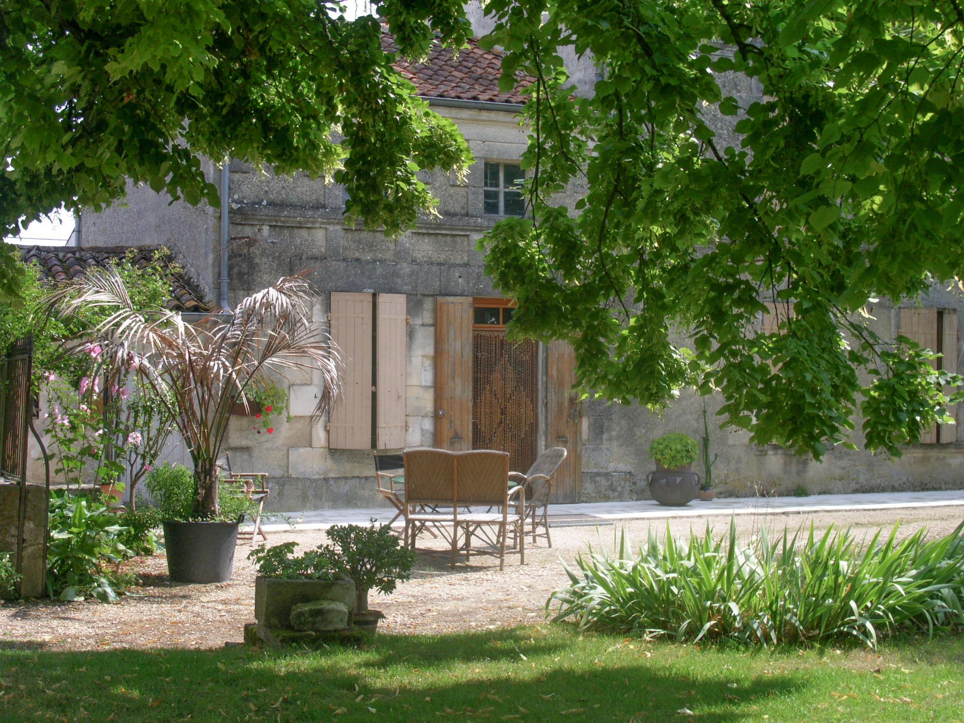 - Gérard Vaubourg maison charentaise en pierre visible à travers le feuillage d'un énorme tilleur au premier plan, mobilier de jardin en rotin et palmier dans un pot situé à gauche de l'image, massif d'iris à droite